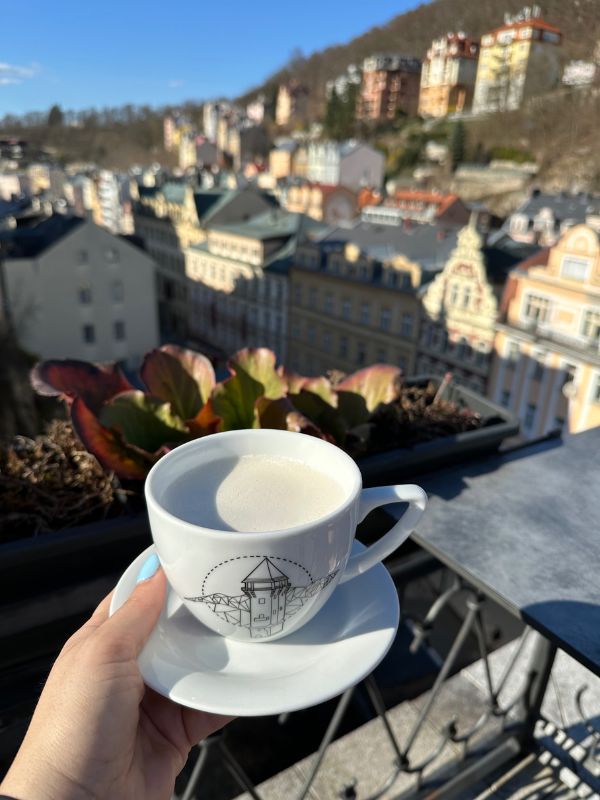 Hrníček s kávou držený nad budovami Karlových Varů.