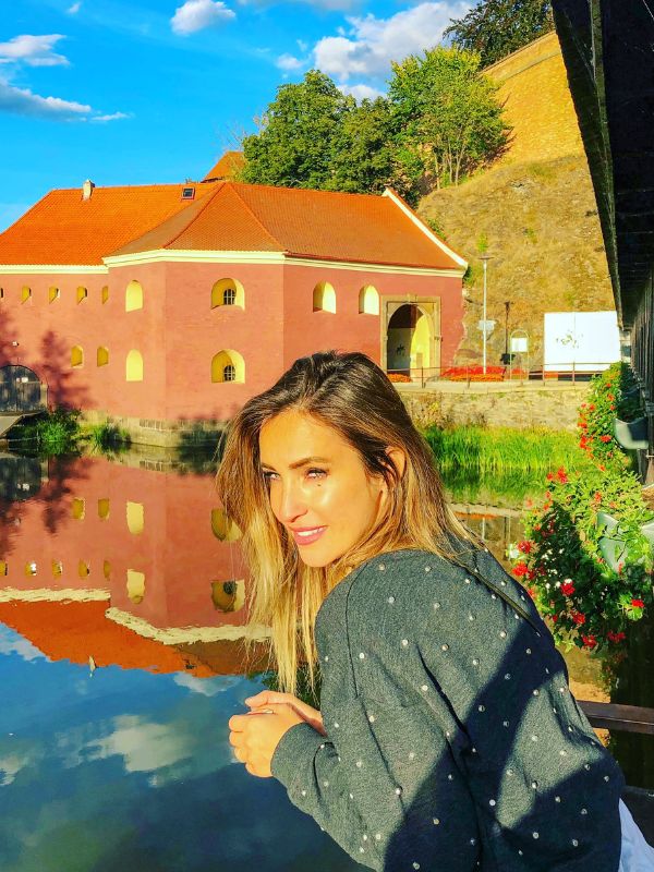 Žena pózuje před oranžovou budovou s odrazem ve vodě pod modrou oblohou.