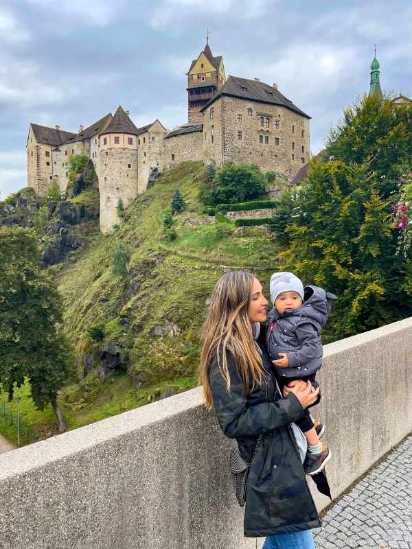 Žena drží malého chlapce s hradem Loket na zeleném kopci v pozadí.