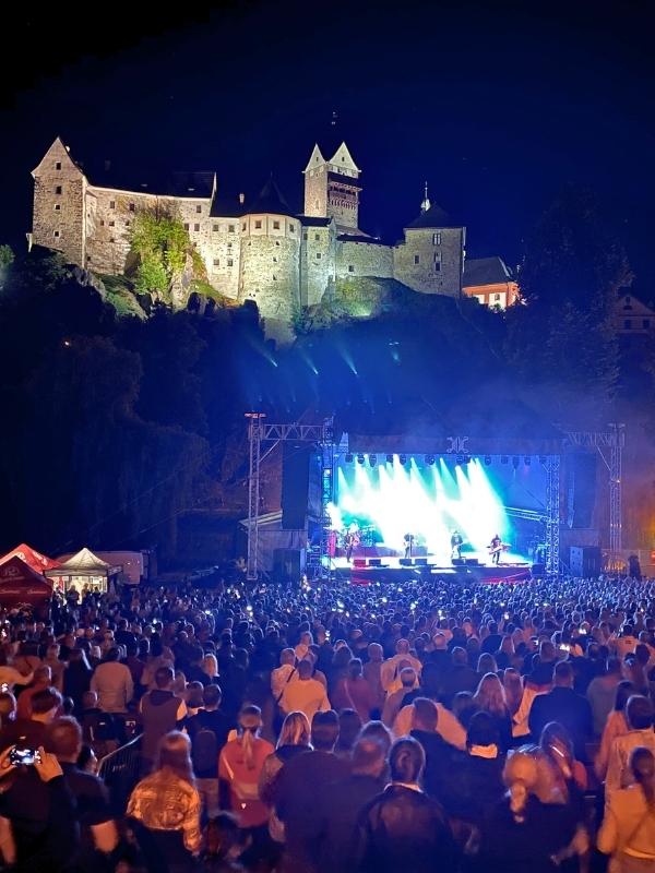 Osvětlený hrad Loket v noci s koncertem a stovkami lidí v popředí.