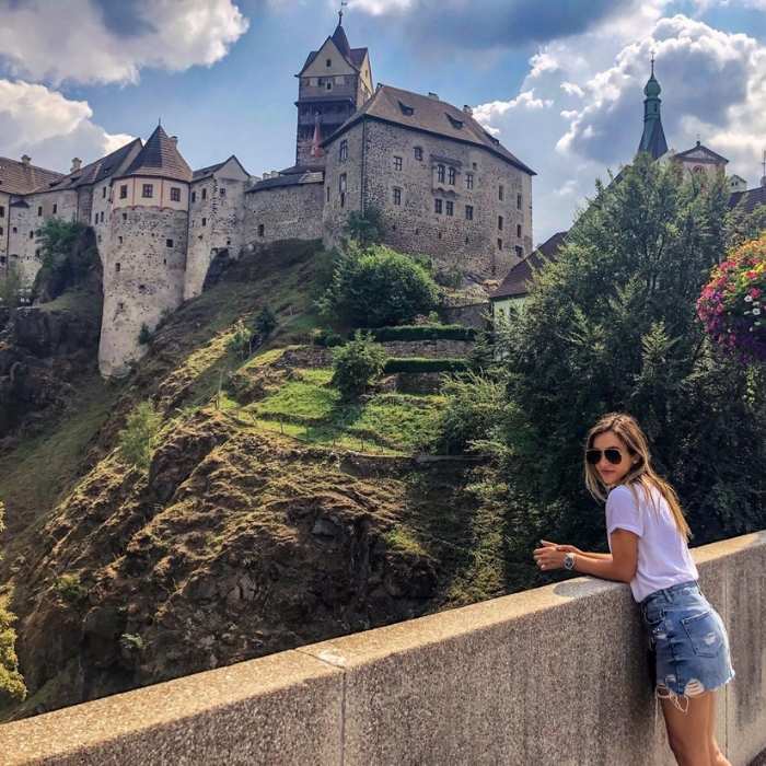 Žena ve slunečních brýlích pózuje před hradem Loket na zeleném kopci.