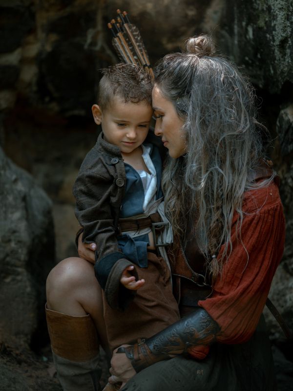 Žena ve starověkém obleku s šípy na zádech a vlasy staženými do drdolu objímá svého malého syna