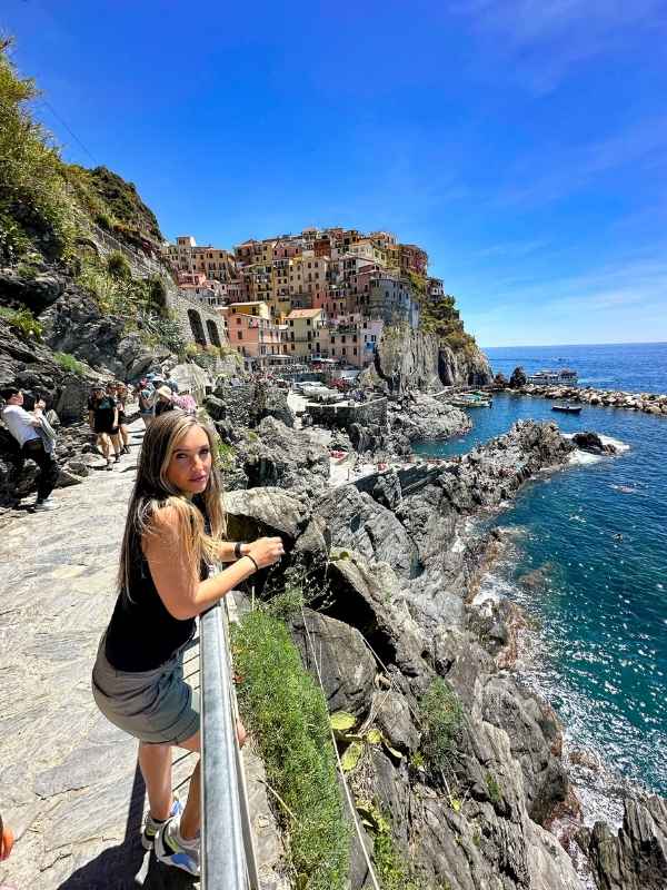 žena se opírá o zábradlí nad oceánem, v pozadí útes s městečkem Corniglia