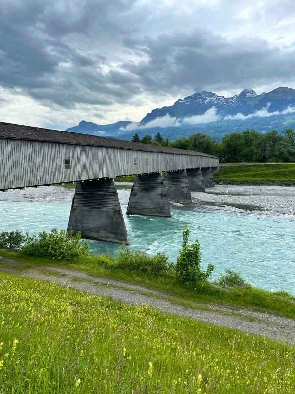 Dřevěný most mezi Lichtenštejnskem a Švýcarskem nad tyrkysově modrou řekou obklopenou zelení.