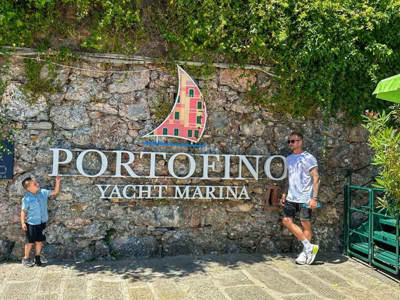 muž s tetováním a chlapec sedí na molu v Portofinu, zelený kopec s barevnými domky v pozadí