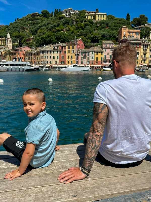 muž s tetováním a chlapec sedí na molu v Portofinu, zelený kopec s barevnými domky v pozadí