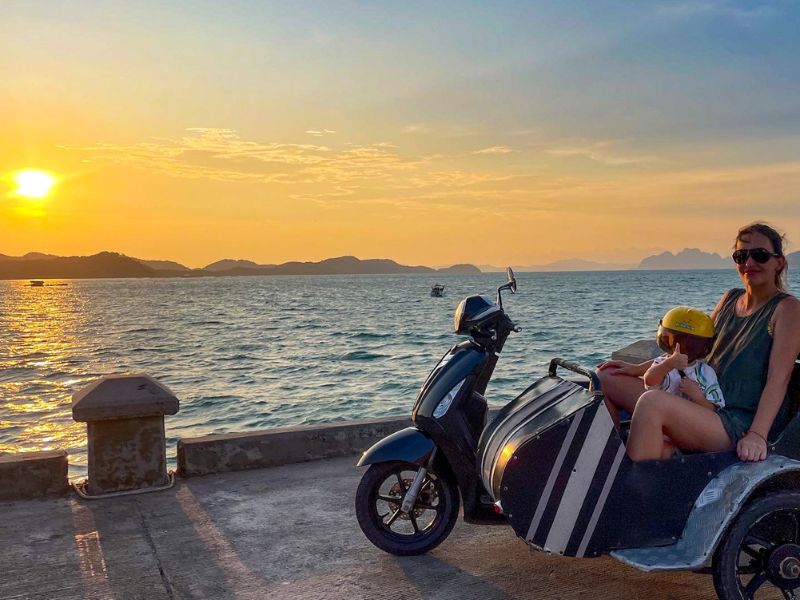 Žena sedí s dítětem na motorce se sidecarem, za nimi se rozprostírá oceán.