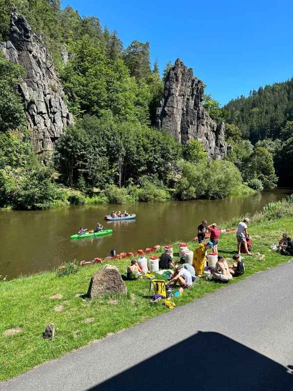 Lidé sedí na trávě a sjíždějí řeku Ohři v kánoích kolem svatošských skal.