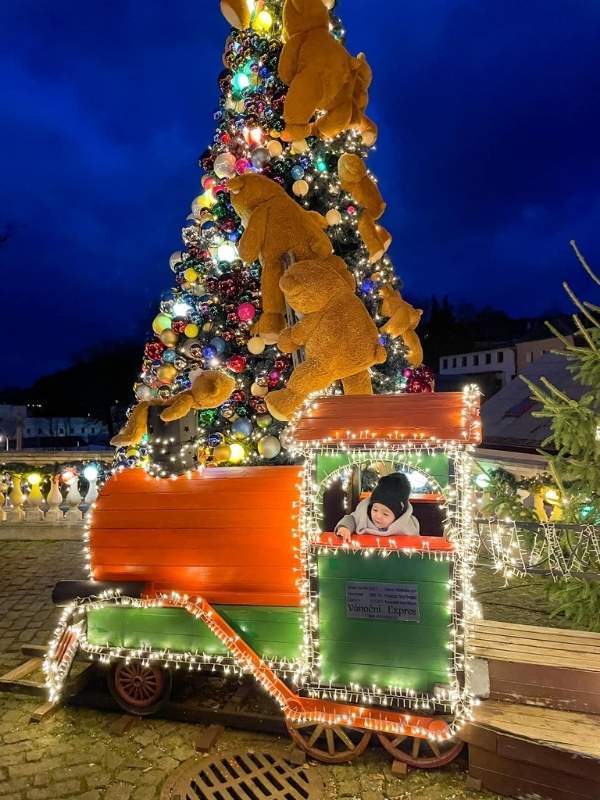 Chlapec kouká z dětského vláčku, za ním velký ozdobený vánoční stromeček, po kterém lezou plyšoví medvědi.