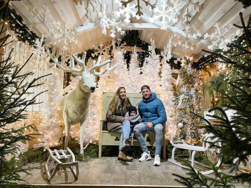 Mladá rodina sedí na lavičce obklopena zasněženými stromy a bílým sobem, saně a umělý sníh dodávají vánoční atmosféru.