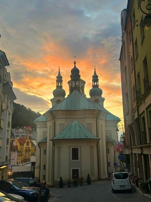 Kostel sv. Máří Magdaleny v Karlových Varech při západu slunce – barokní kostel s typickou zelenou měděnou střechou a věžičkami, obklopený městskými domy, za nímž se rozprostírá barevná obloha.