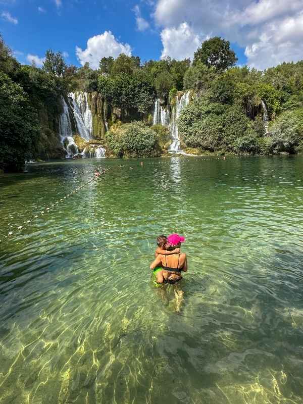 Kravice Bosna a Hercegovina **Alt text:** Žena stojí ve vodě a drží malé dítě v náručí, oba jsou otočeni zády k fotoaparátu. Stojí v průzračně zelené laguně pod vodopády Kravica v Bosně a Hercegovině. Kolem je bujná zeleň a několik kaskád padající vody, na obloze jsou bílé mraky a modré nebe. Atmosféra je klidná a krásná, jako moment rodinné pohody uprostřed přírody.