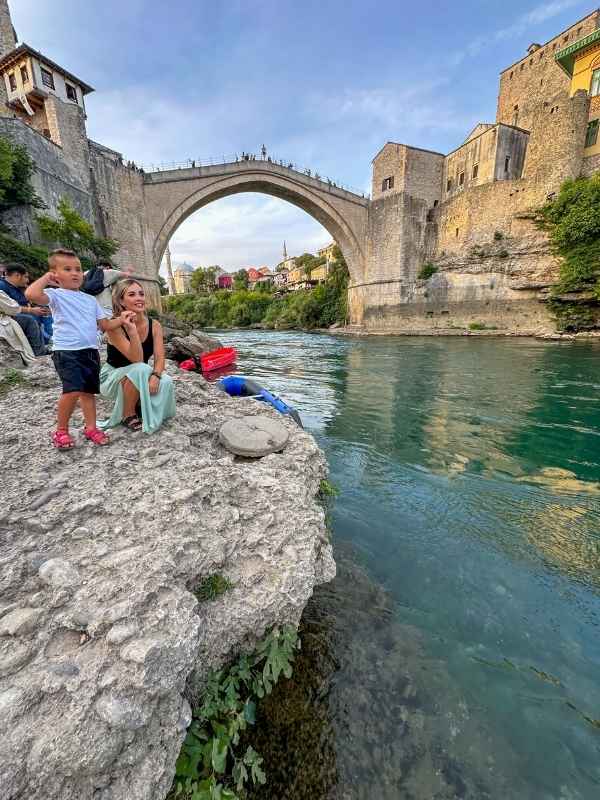 Mostar Bosna Žena sedí na skalnatém břehu řeky Neretvy pod ikonickým Starým mostem v Mostaru, vedle ní stojí malé dítě. Za nimi se klene historický kamenný oblouk mostu a staré městské budovy. Voda pod nimi je průzračně tyrkysová a odráží večerní světlo. Scéna působí uvolněně a cestovatelsky, jako poklidný moment obdivu k jednomu z nejznámějších symbolů Bosny a Hercegoviny.