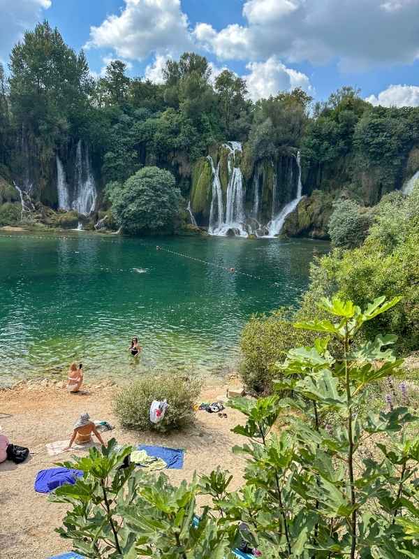 Vodopády Kravica **Alt text:** Pohled na vodopády Kravica v Bosně a Hercegovině s tyrkysovou lagunou v popředí. Lidé relaxují na pláži, někteří sedí na ručnících, jiní se koupou ve vodě. Vodopády padají z několikametrové výšky přes zelení porostlé skály, kolem je hustá vegetace a nad nimi modré nebe s bílými mraky. Atmosféra připomíná přírodní koupaliště uprostřed tropického ráje.