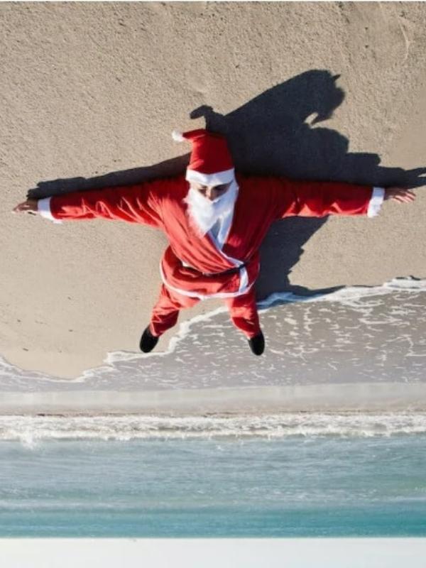Santa na plazi Letecký pohled na úzký červený most nad klidnou řekou, obklopený bujnou zelení a stromy v podzimních barvách. Dva lidé po něm kráčí, zachycujíce poklidný okamžik spojený s přírodou.